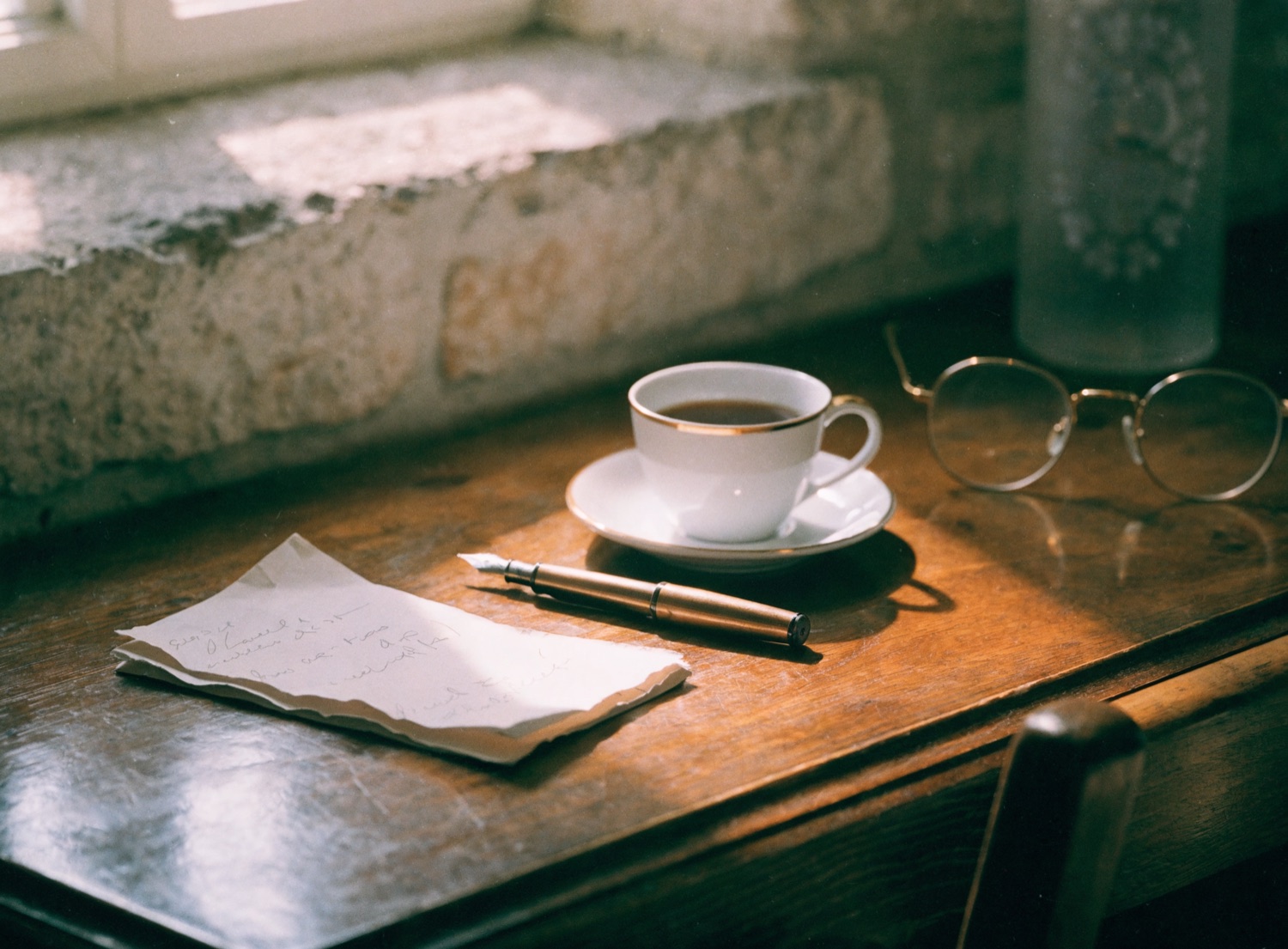 A fountain pen, a handwritten letter, a cup of tea and reading glasses on a timber desk in soft light